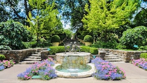 Um jardim com uma fonte central, degraus de pedra e canteiros de flores.