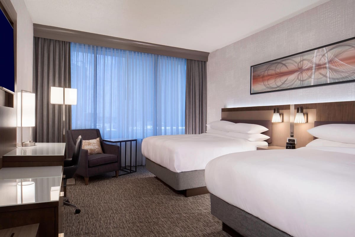 A hotel room with two beds, a desk, a chair, and a large window with curtains.