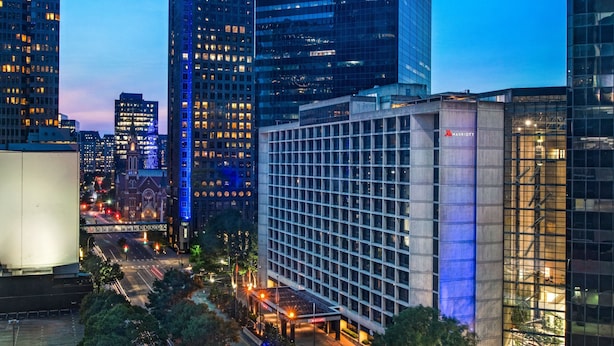 Um hotel moderno com fachada de vidro, rodeado por edifícios altos e uma rua movimentada ao entardecer.