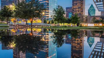 A cityscape with modern high-rise buildings reflected in a calm body of water, surrounded by trees and a pedestrian bridge.