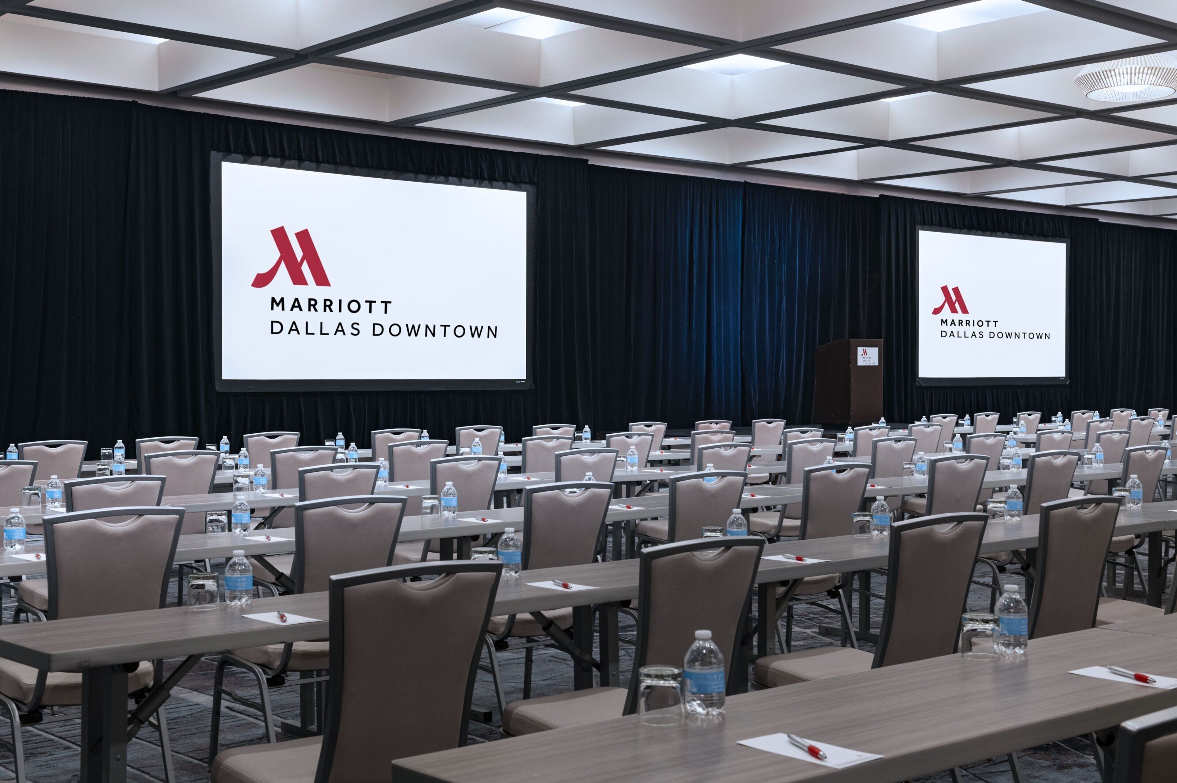 Un salón de conferencias con filas de mesas, sillas y pantallas grandes mostrando el logotipo de Marriott Dallas Downtown.