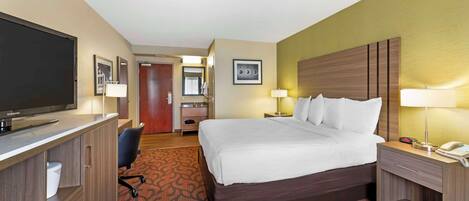 A hotel room with a bed, bedside table, lamp, desk with a television, and a carpeted floor.