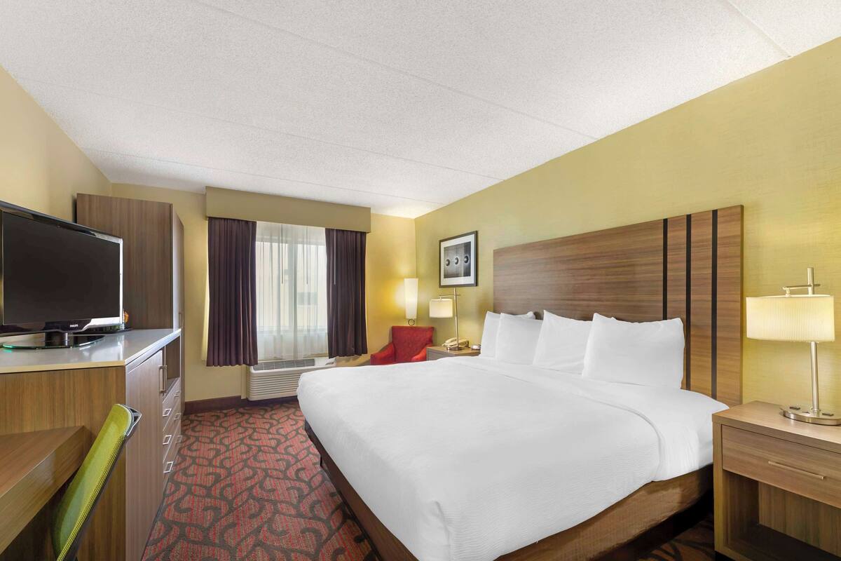 A hotel room with a large bed, a desk with a television, a red chair, a nightstand with a lamp, and a window with curtains.