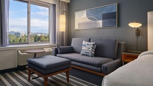 Une chambre d’hôtel moderne avec un canapé gris, une petite table, un lampadaire et une grande fenêtre donnant sur la ville.