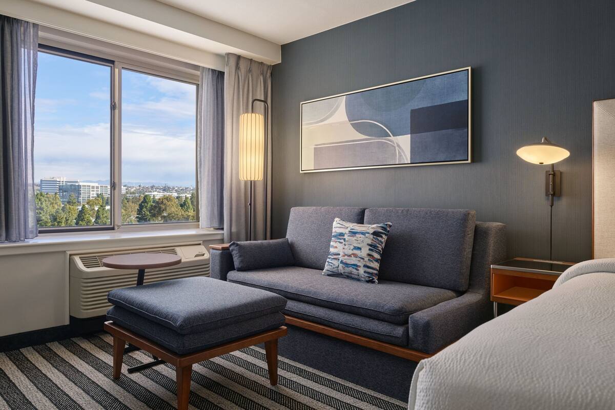 A modern hotel room with a grey sofa, a small table, a standing lamp, and a large window offering a city view.