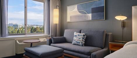 Habitación de hotel moderna con sofá gris, una mesita, una lámpara de pie y un amplio ventanal con vistas a la ciudad.