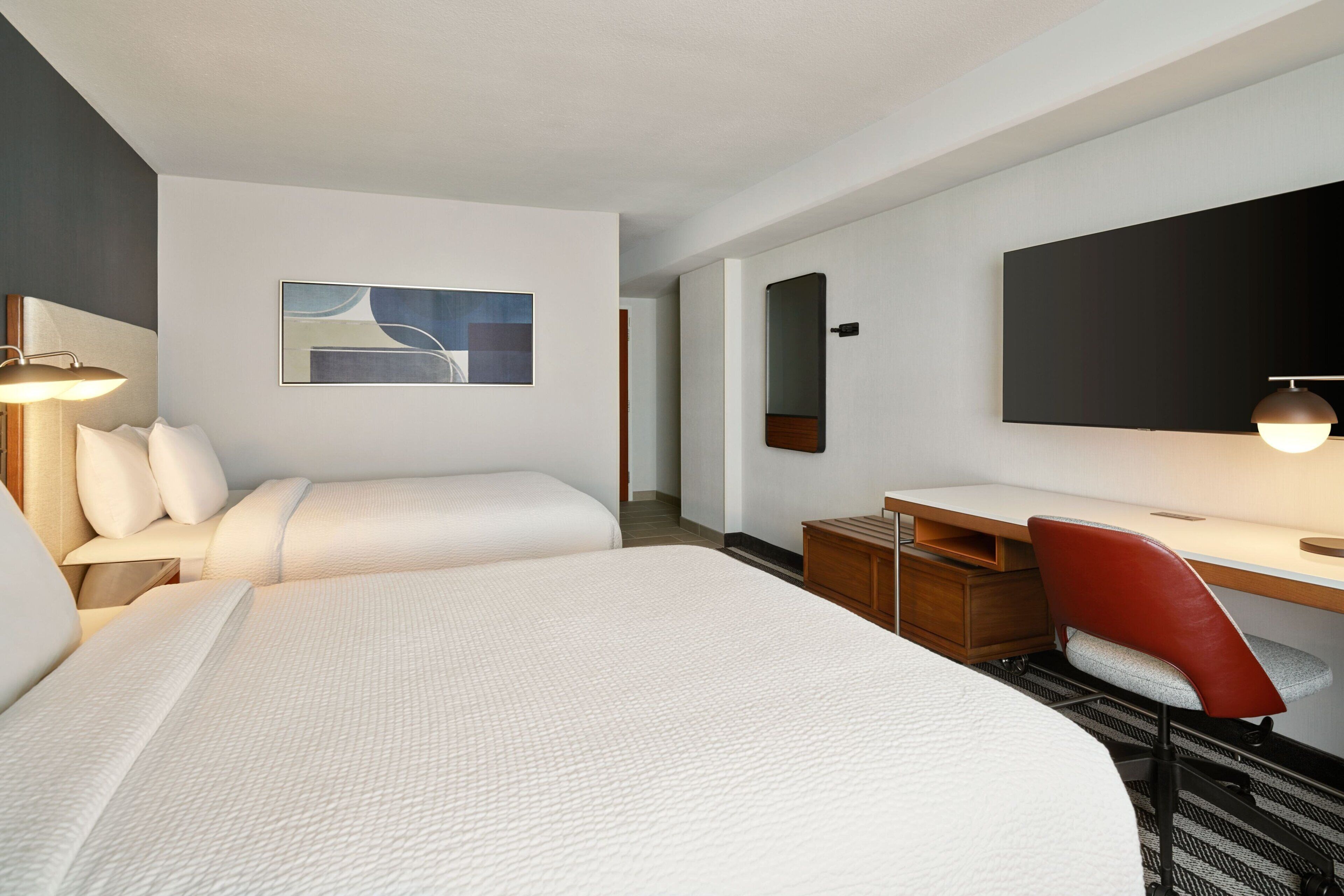 Une chambre d’hôtel avec deux lits, un bureau, une chaise, un téléviseur à écran plat et un tableau au mur.