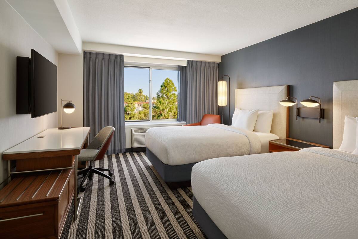 A hotel room with two beds, a desk, a chair, a flat-screen TV, and a window with curtains.