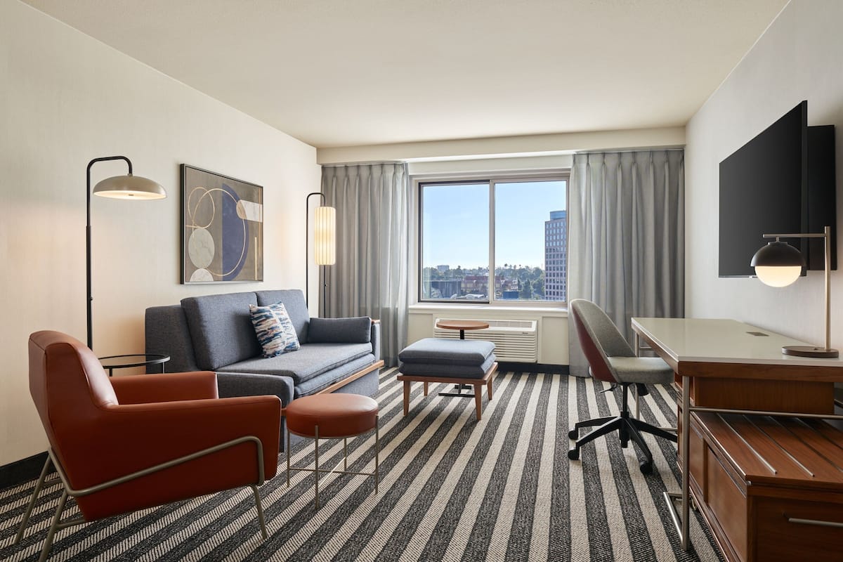 A hotel room with a sofa, armchair, ottoman, desk, and a large window with a city view.