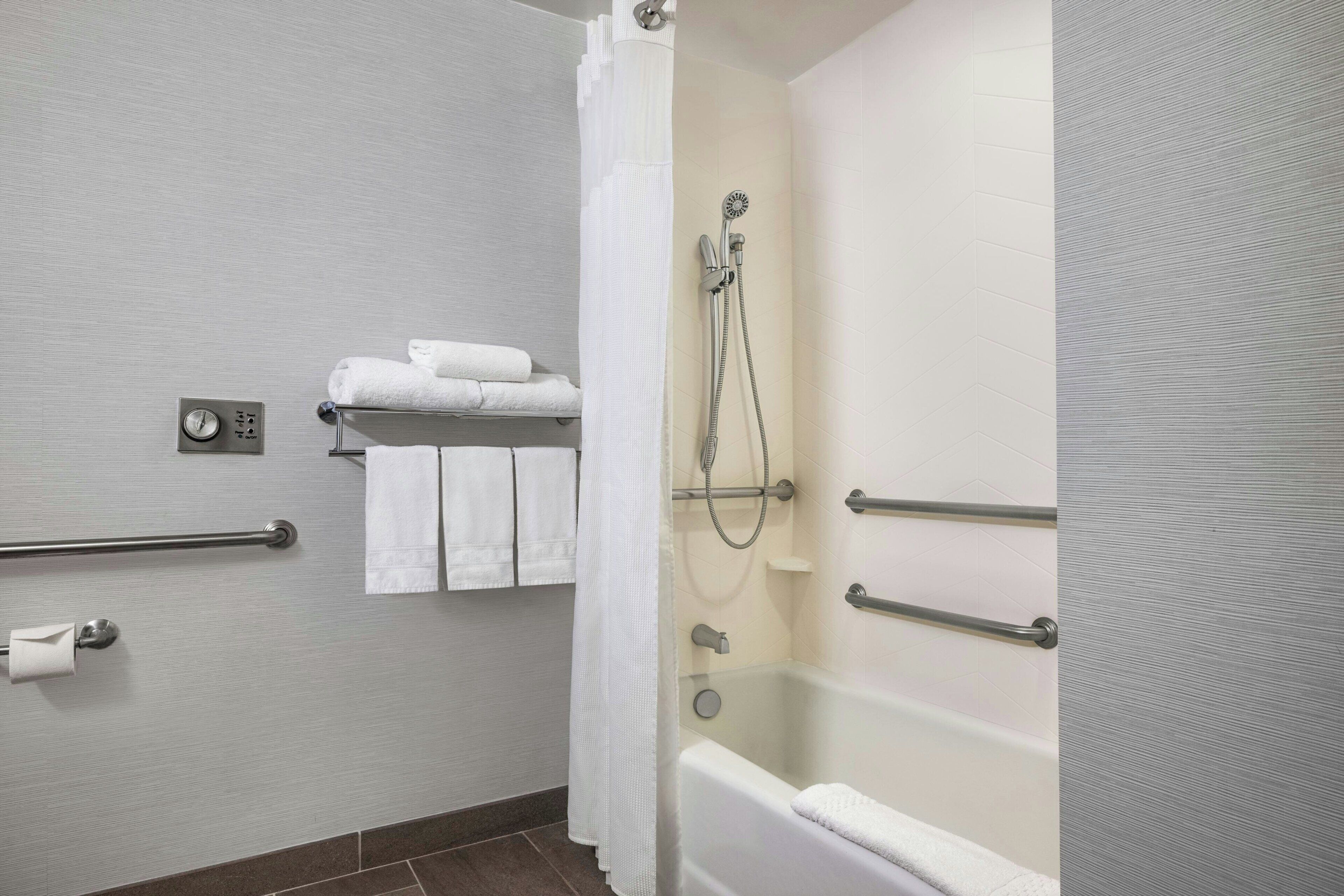 Un bagno con una vasca da bagno, una doccia, un portasciugamani con asciugamani piegati, un porta carta igienica e una maniglia di sostegno.