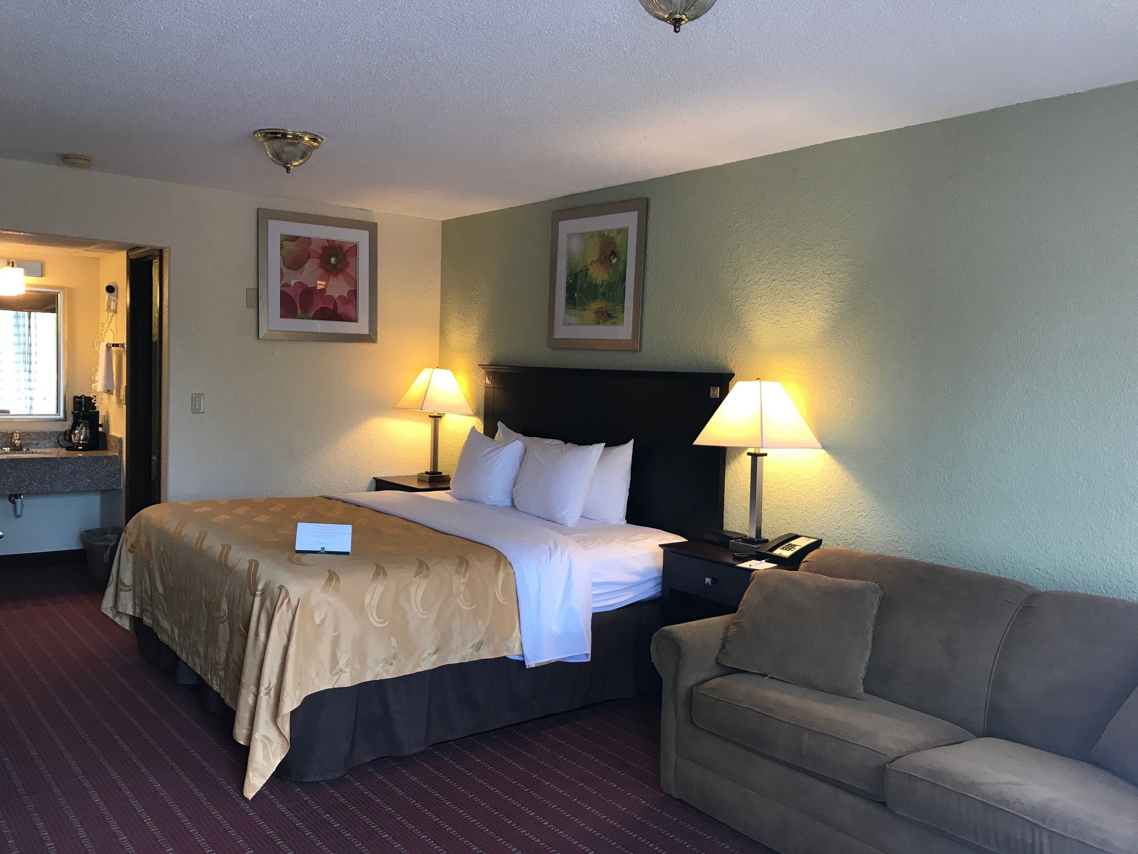 A hotel room with a bed, two bedside lamps, a nightstand, a sofa, and a painting on the wall.