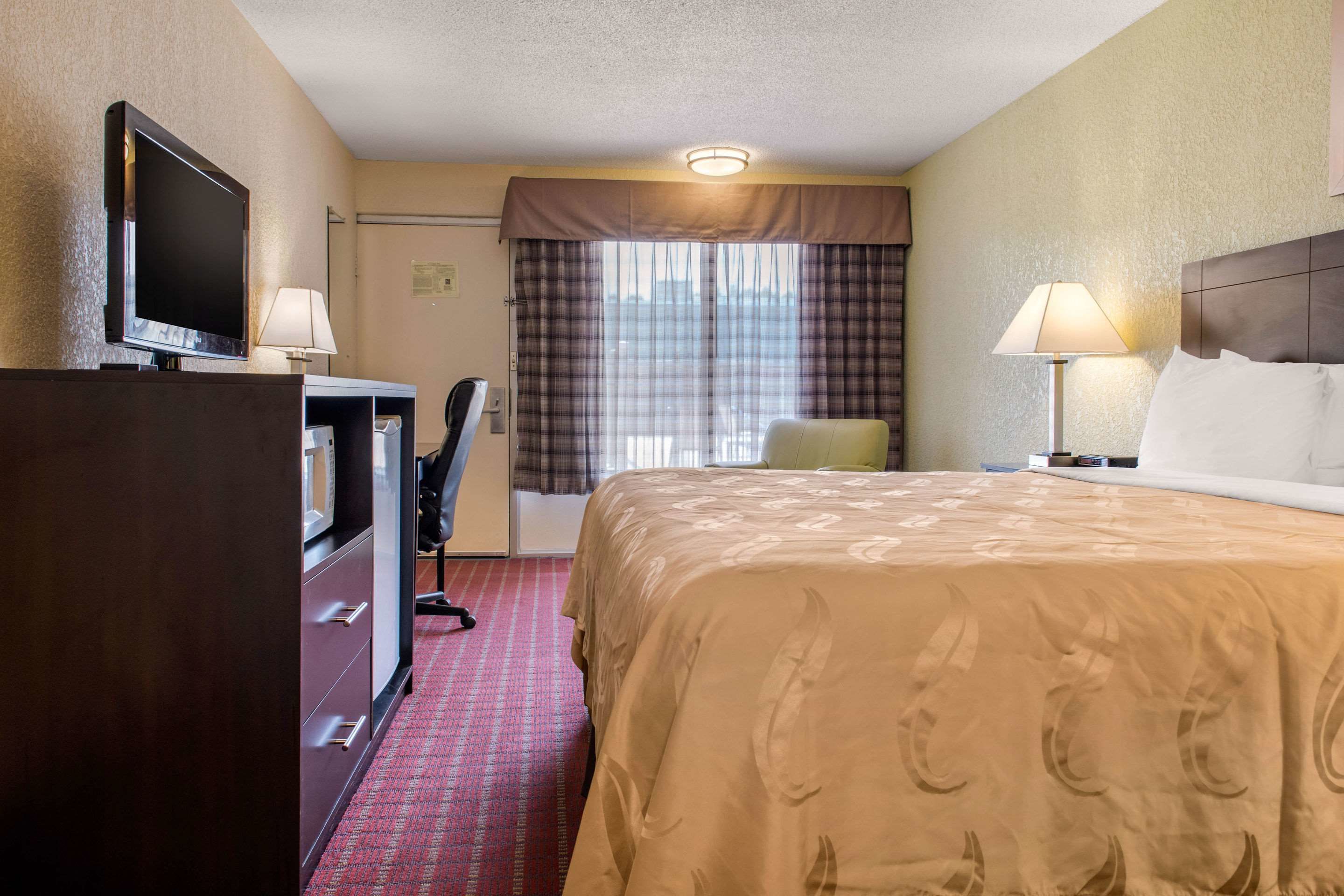 Une chambre d’hôtel avec un lit, un bureau avec un ordinateur, une télévision et une fenêtre avec des rideaux.