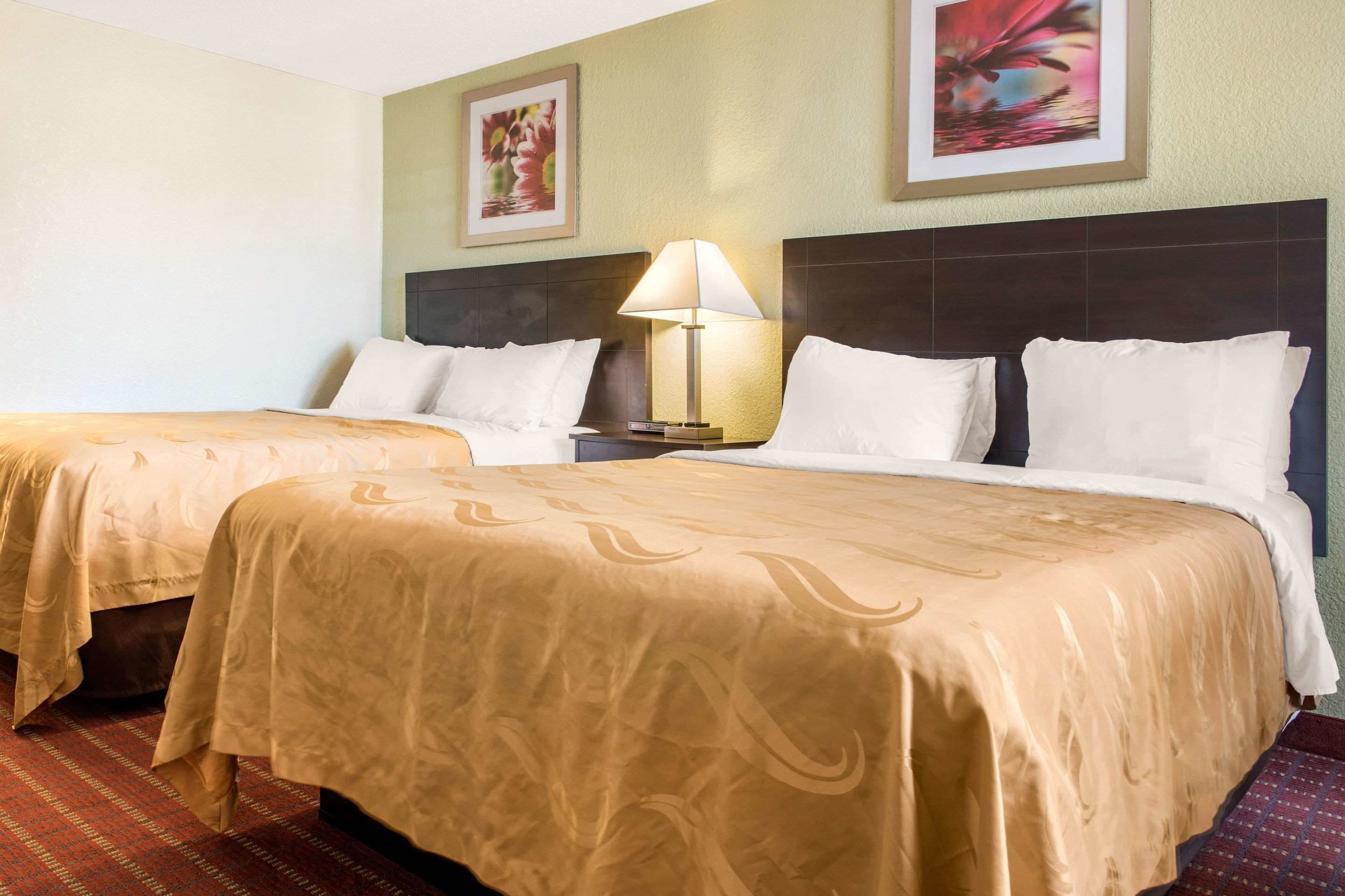 A hotel room with two beds, each with a beige bedspread and white pillows. There is a nightstand with a lamp between the beds. The walls are painted in a light color, and there are two framed pictures above the beds.