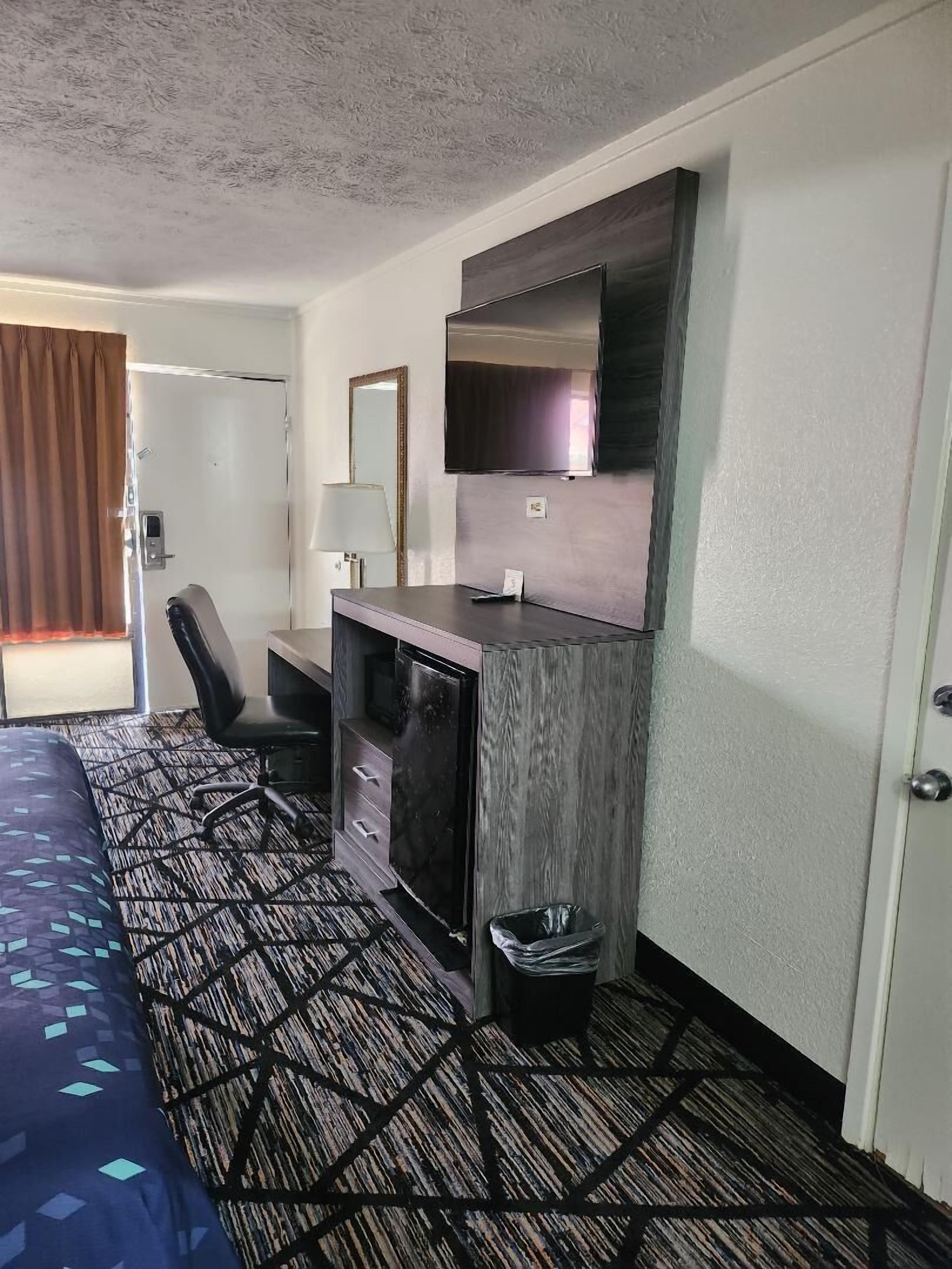 A hotel room with a bed, a desk with a TV, a chair, a lamp, and a trash bin.