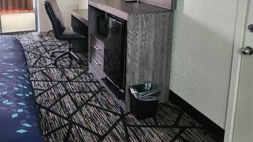 A hotel room with a bed, a desk with a TV, a chair, a lamp, and a trash bin.