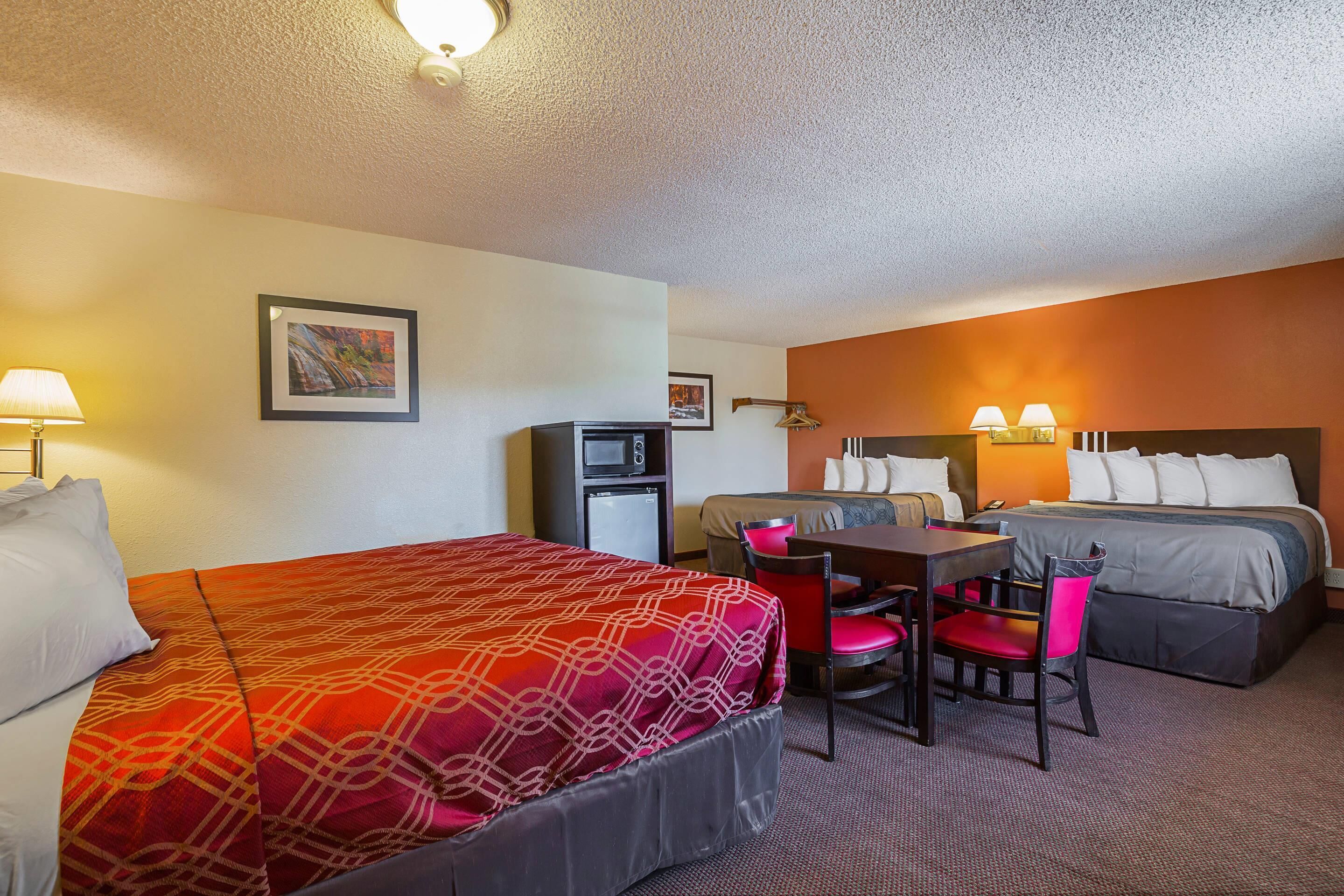 A hotel room with two beds, a desk, and chairs. There is a refrigerator and a lamp on the desk.