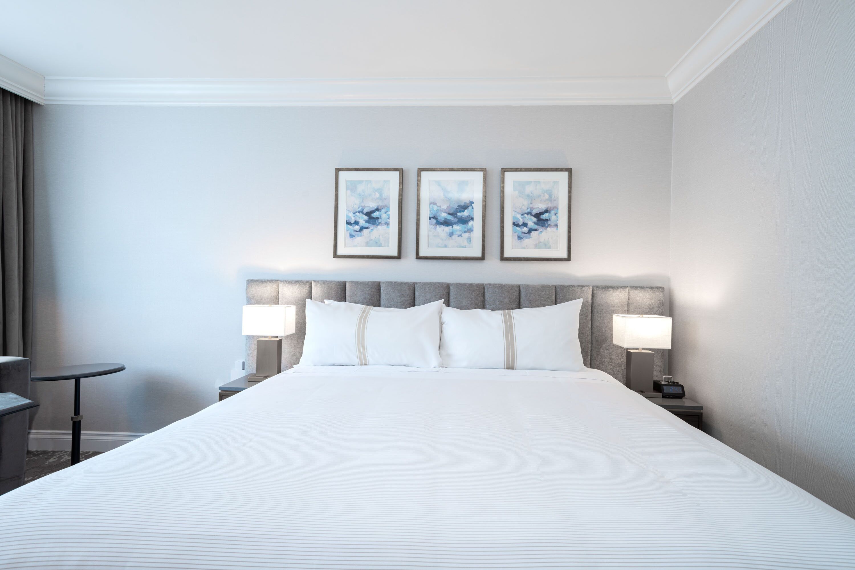 A hotel room with a large bed, two bedside tables with lamps, a grey upholstered headboard, and three framed artworks on the wall.