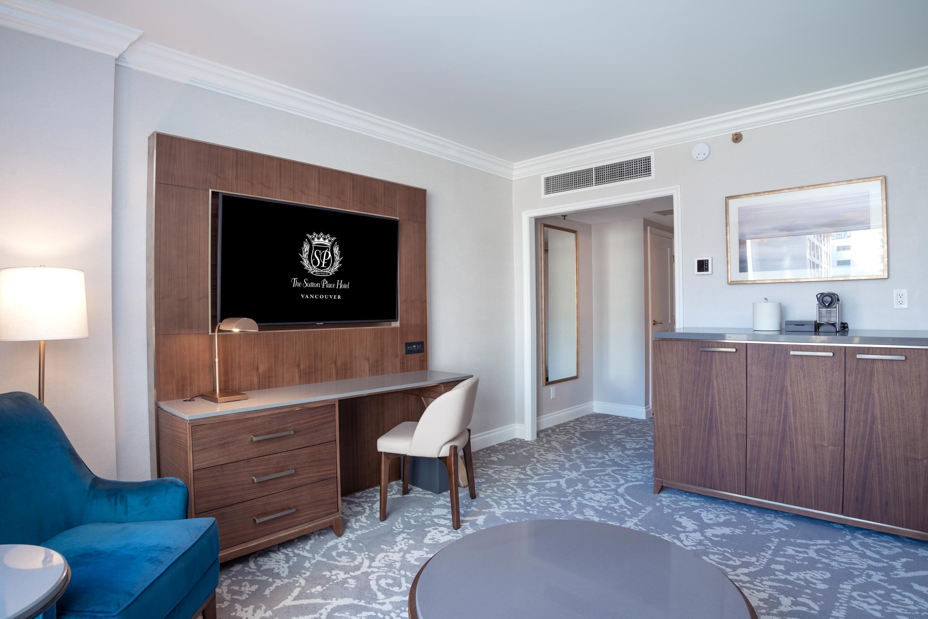 A hotel room with a flat-screen TV mounted on a wooden cabinet, a desk with a chair, a round table, a minibar, and a large mirror on the wall.