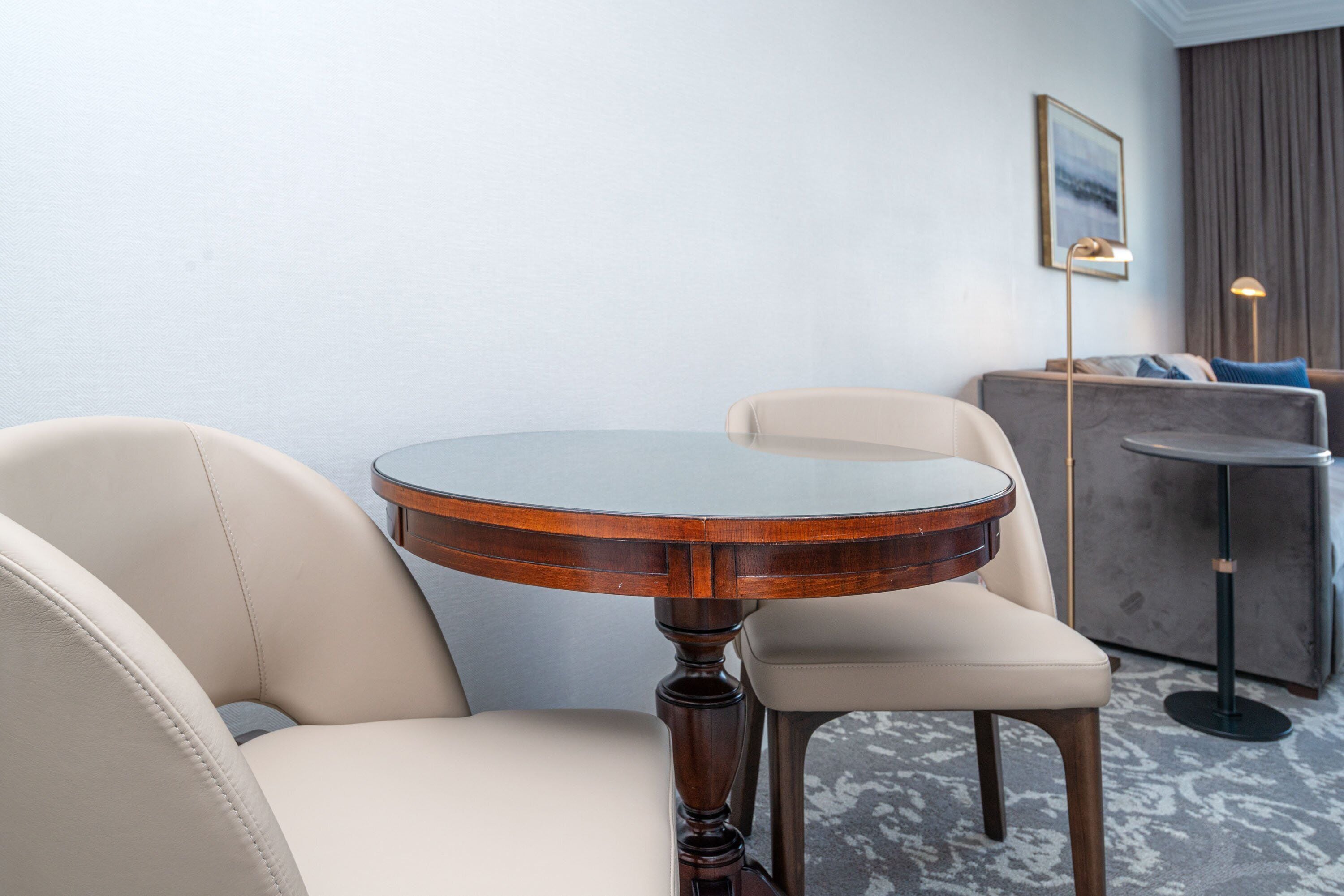 A round wooden table with a glass top, two beige chairs, a grey sofa, and a floor lamp in a room with a light blue wall and a framed picture.