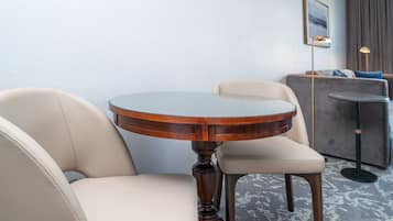 A round wooden table with a glass top, two beige chairs, a grey sofa, and a floor lamp in a room with a light blue wall and a framed picture.