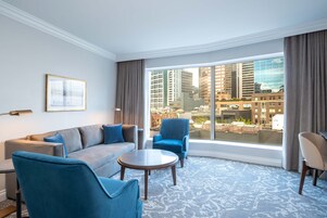 A modern hotel room with a large window, a sofa, two armchairs, and a small table.
