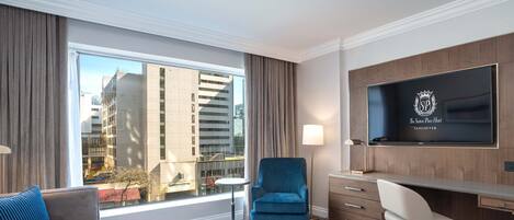 A hotel room with a large window, a sofa, a round table, a blue armchair, a desk with a chair, and a flat-screen TV displaying the hotel logo.