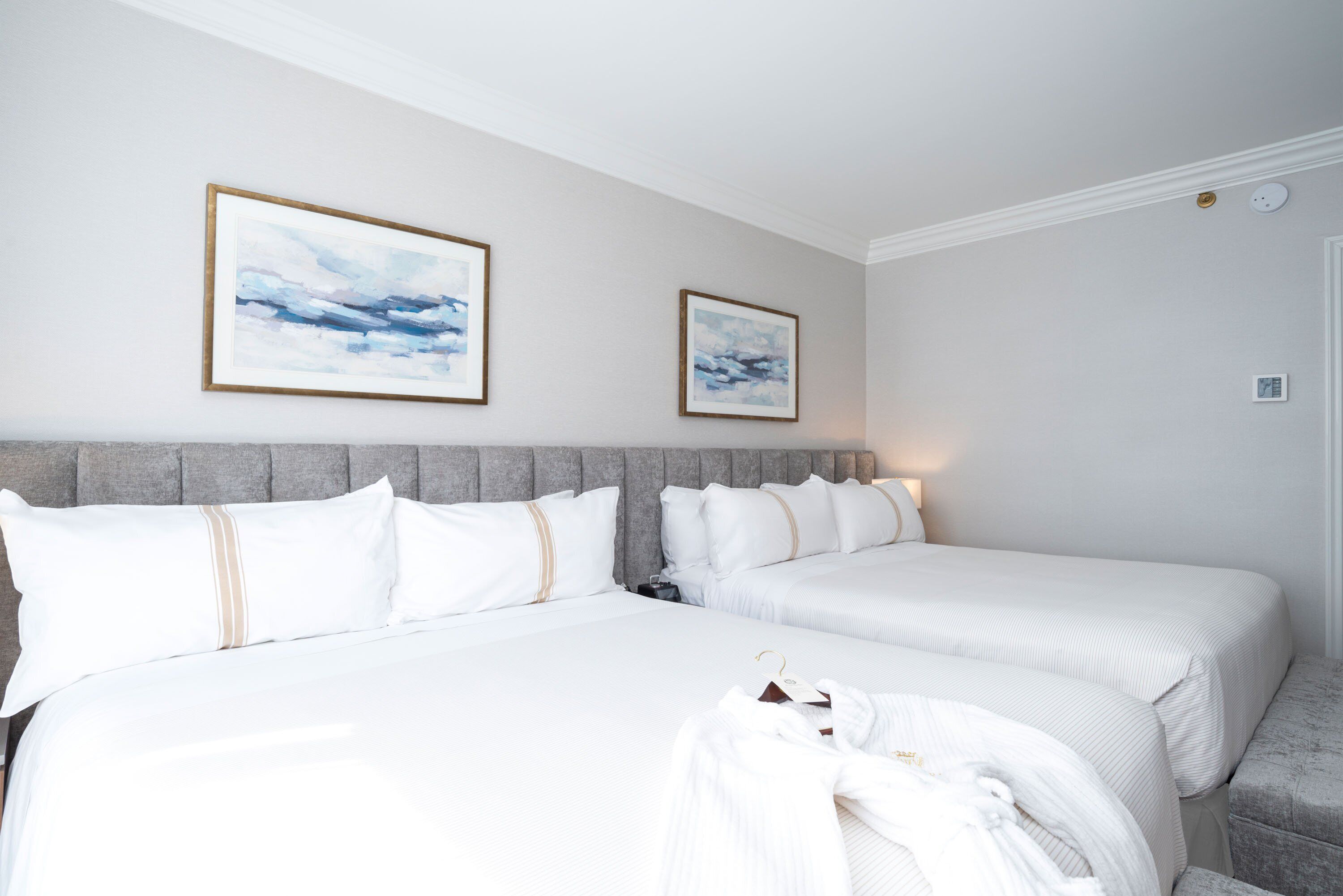 Una habitación de hotel con dos camas, cabecera gris, ropa de cama blanca y dos cuadros en la pared.