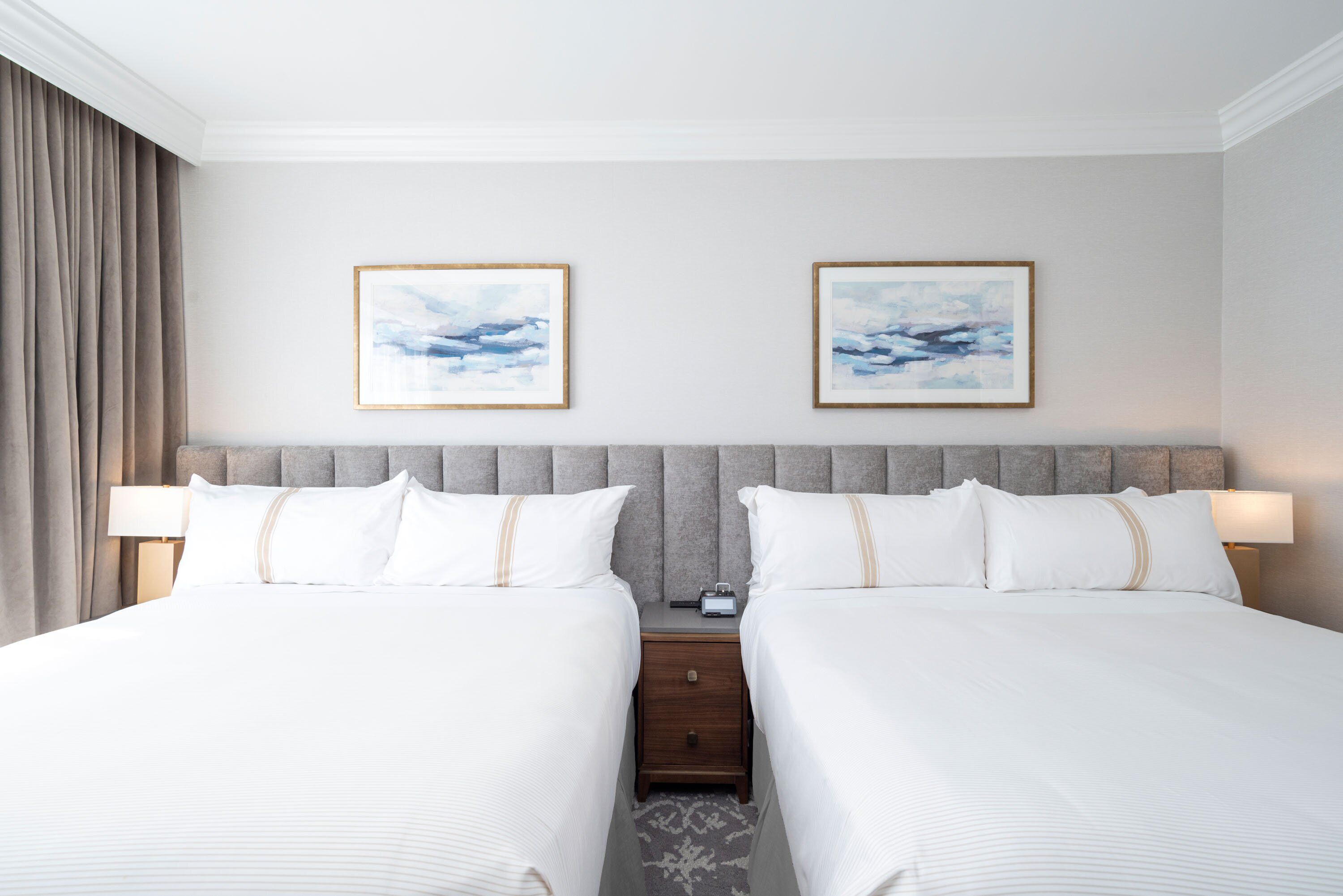 Une chambre d’hôtel avec deux lits, une tête de lit grise en tissu, du linge de lit blanc et deux tableaux abstraits encadrés accrochés au mur.