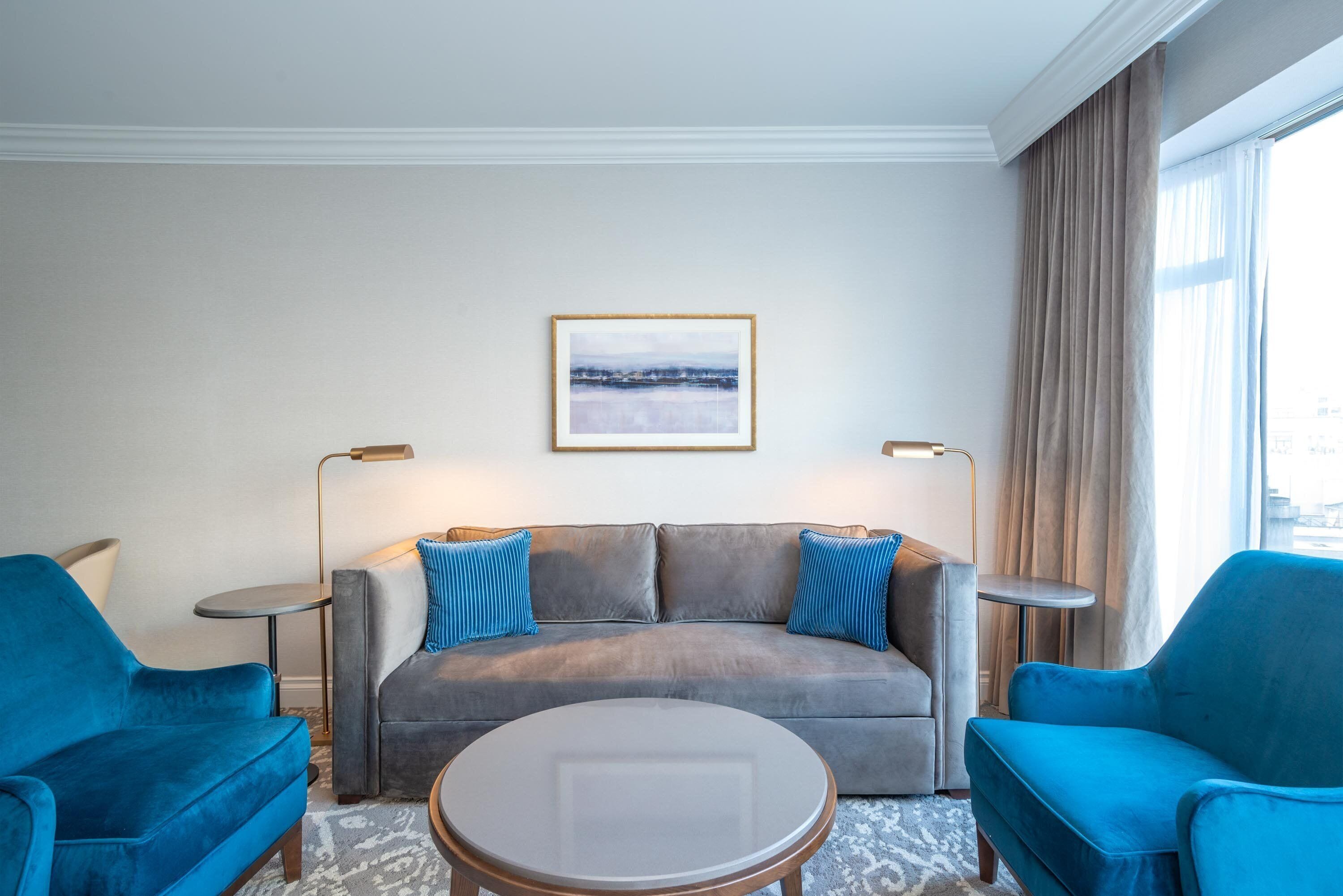 A modern living room with a grey sofa, two blue armchairs, a round coffee table, and a framed picture on the wall.