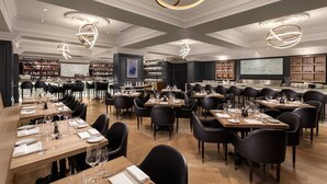 A spacious restaurant with multiple dining tables and black chairs, elegant lighting fixtures, and a bar area with shelves of bottles. - The Sutton Place Hotel Vancouver (Vancouver)