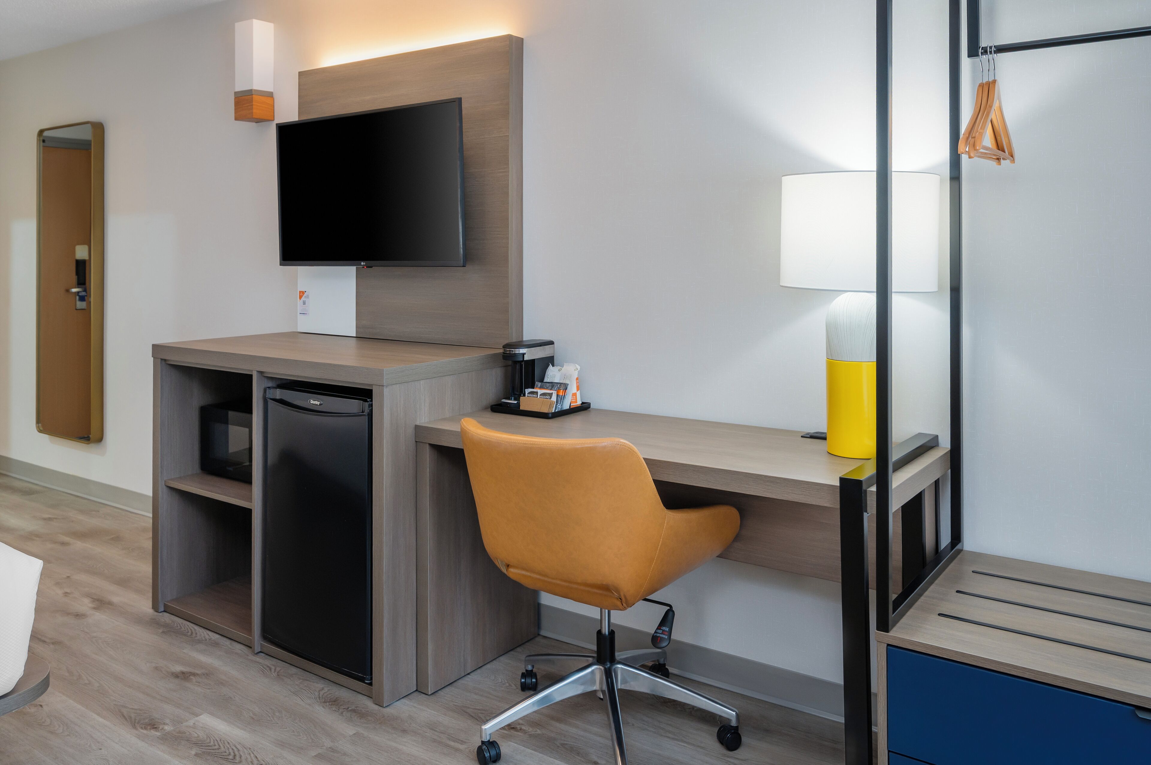 A modern hotel room with a wooden desk, a flat-screen TV, a small refrigerator, a yellow lamp, and a chair.