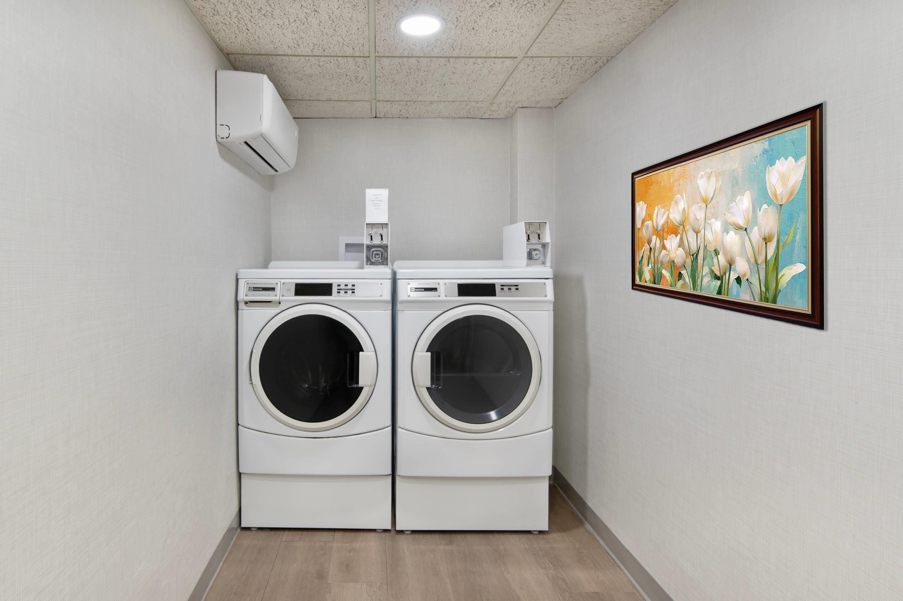 A laundry room with two front-loading washing machines, a wall-mounted air conditioner, and a framed painting of flowers.