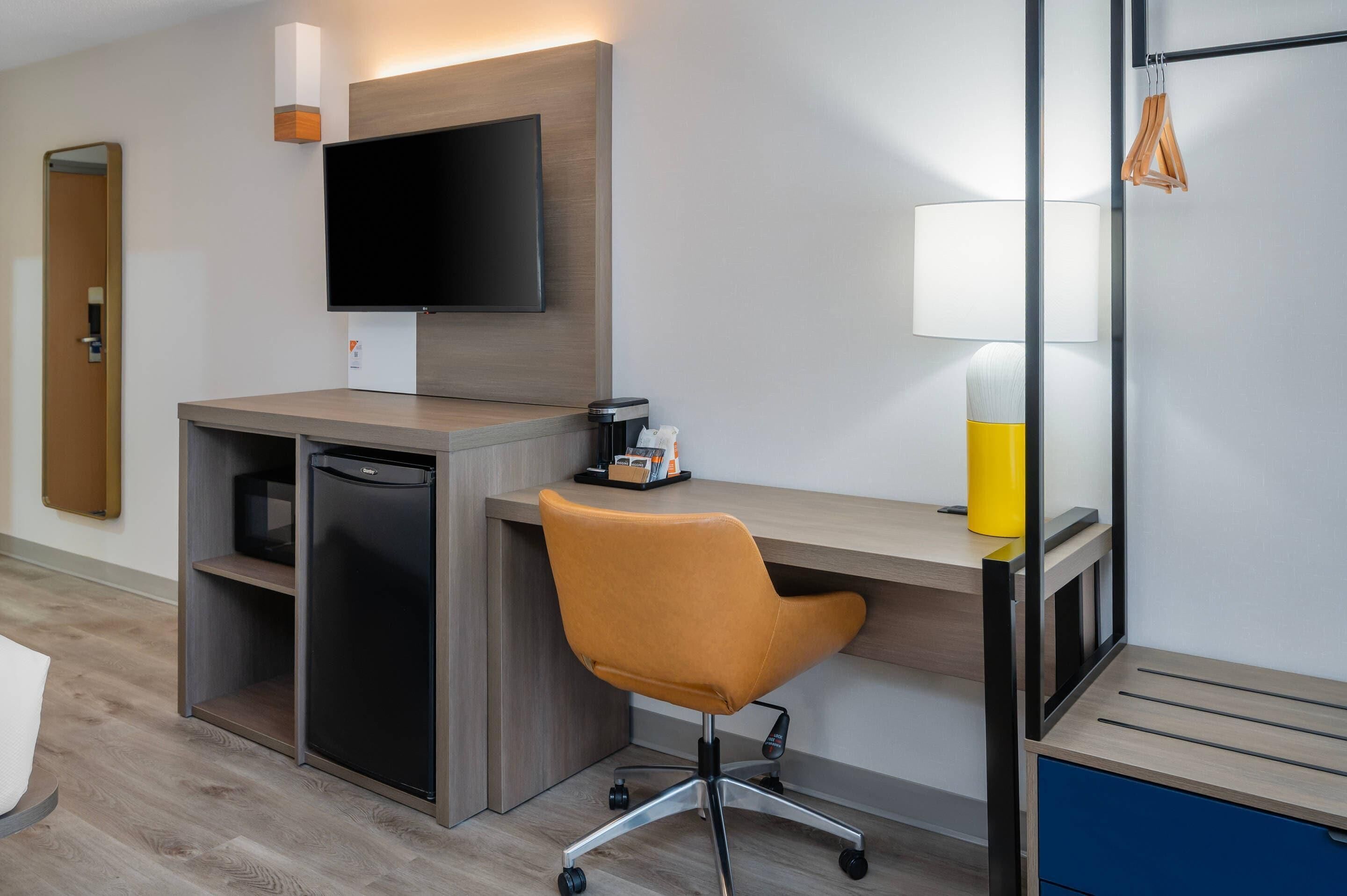 A modern hotel room with a wooden desk, a flat-screen TV, a small refrigerator, a yellow lamp, and a chair.