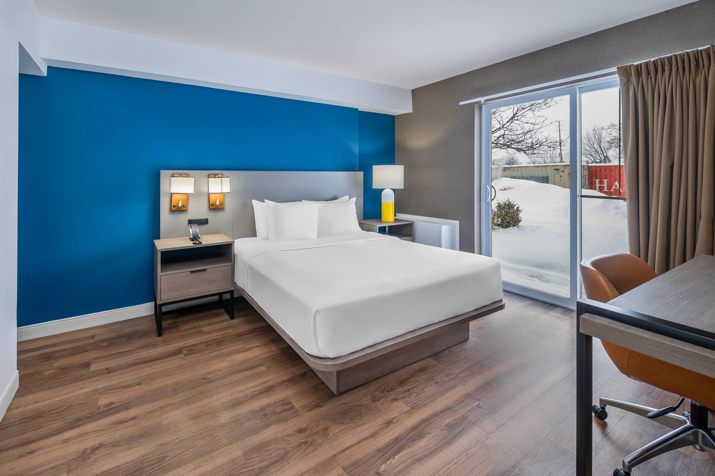 Hotellihuone, jossa on sänky, yöpöytä, lamppu ja liukuovellinen lasiterassi, josta avautuu näkymä lumiseen maisemaan.