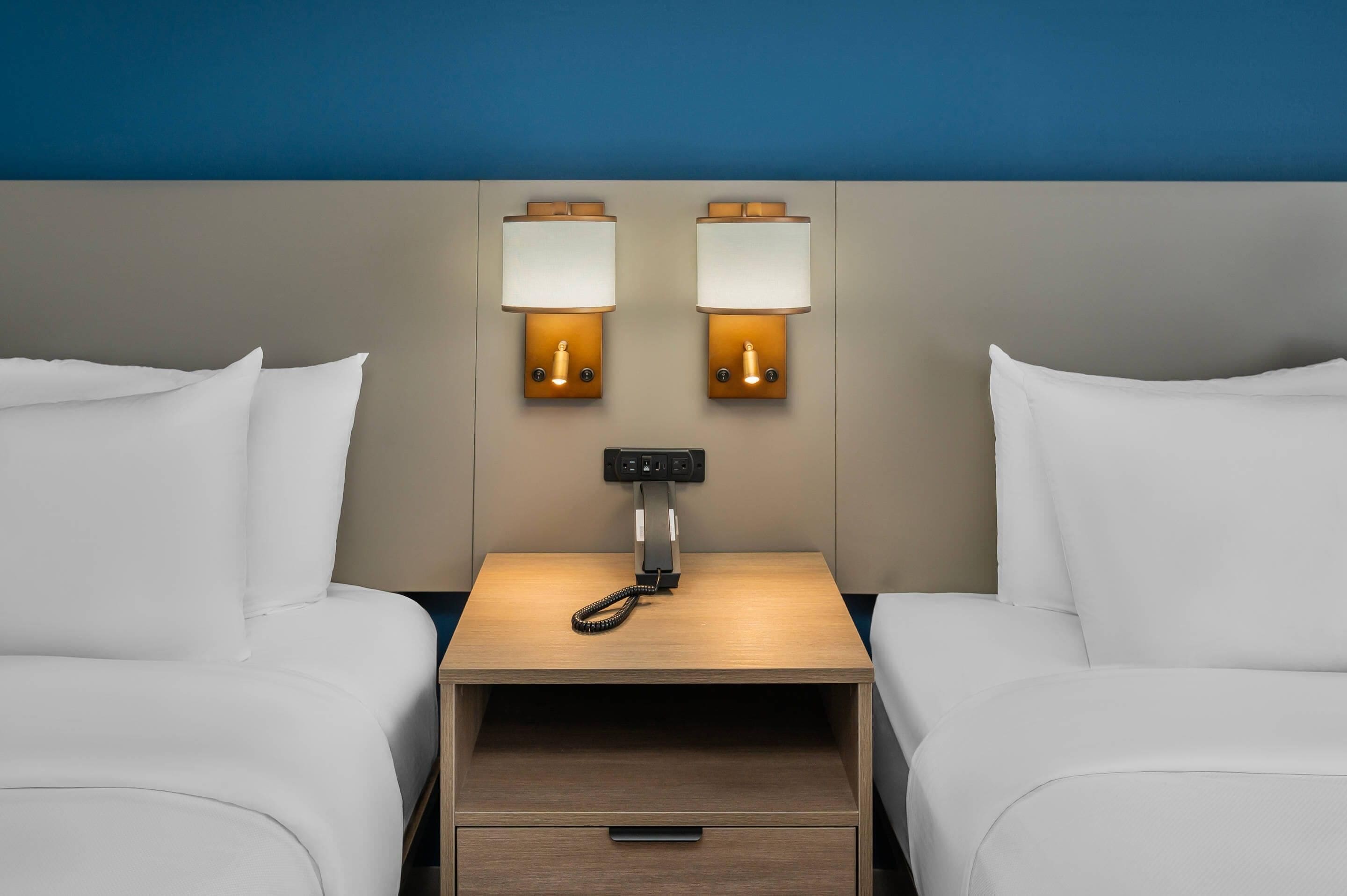 A hotel room with two beds, a nightstand, and two wall-mounted lamps.