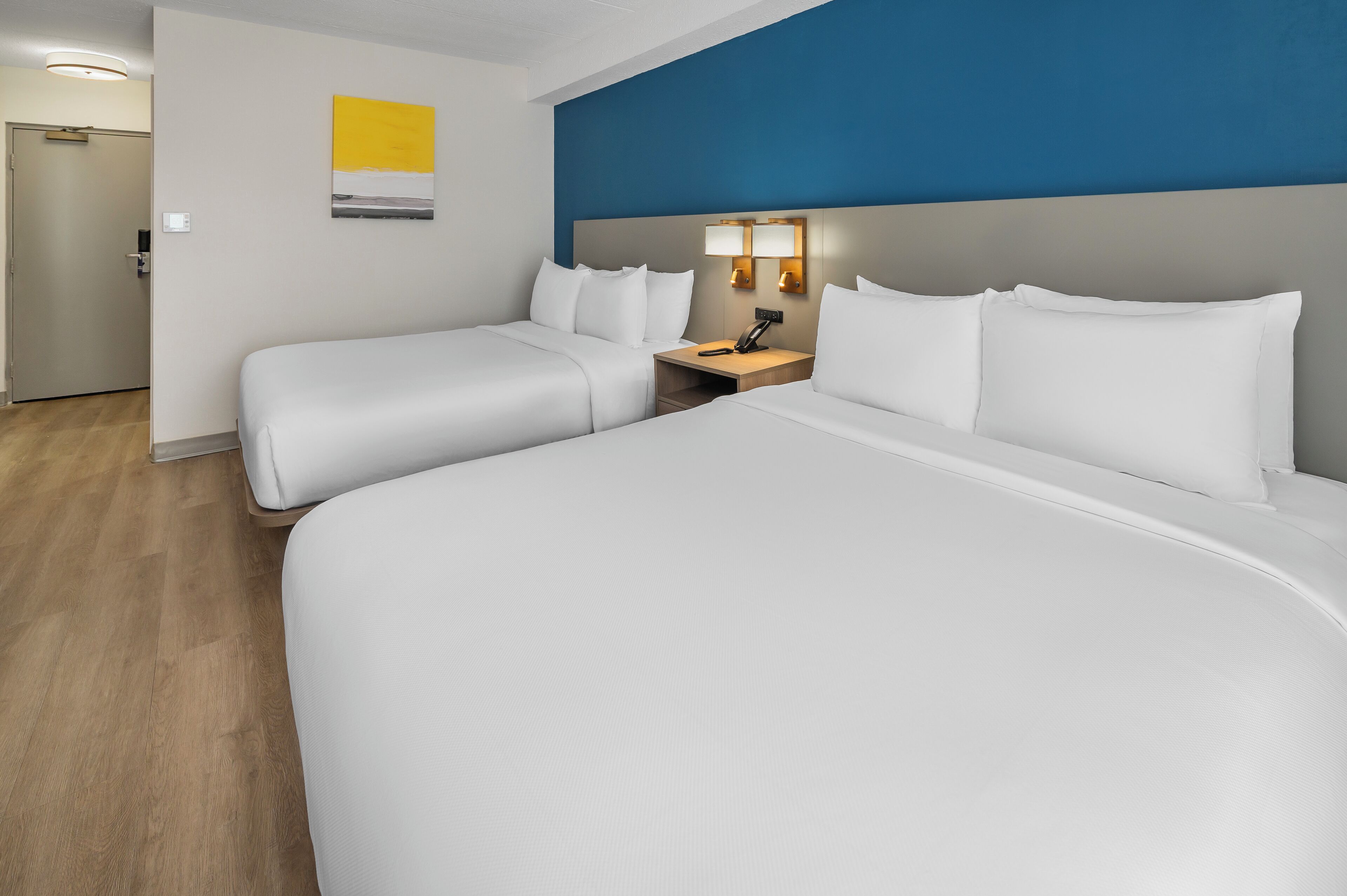 A hotel room with two beds, a nightstand with a lamp, and a door.