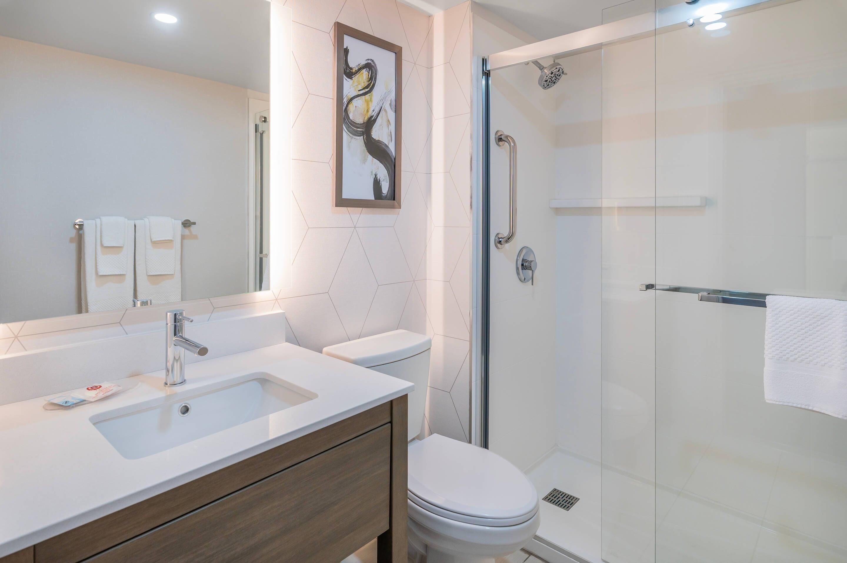 A modern bathroom with a white sink, wooden vanity, a toilet, and a glass-enclosed shower with a handrail.