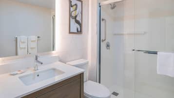 Un baño moderno con un lavamanos blanco, tocador de madera, inodoro y ducha con mampara de vidrio y barra de apoyo.