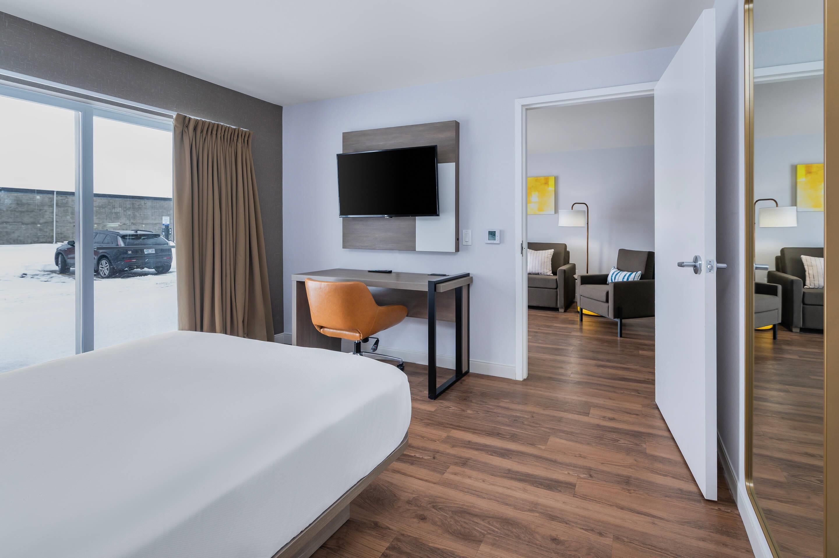 A modern hotel room with a large bed, a desk with a chair, a flat-screen TV, and a view of a parking area.