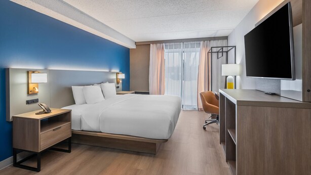 A hotel room with a bed, bedside tables, a desk, a chair, a TV, and curtains.