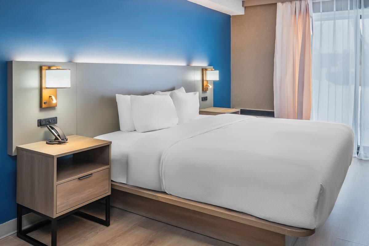 Une chambre d’hôtel moderne, dotée d’un grand lit, de tables de chevet avec des lampes et offrant une vue sur l’extérieur à travers des rideaux légers.