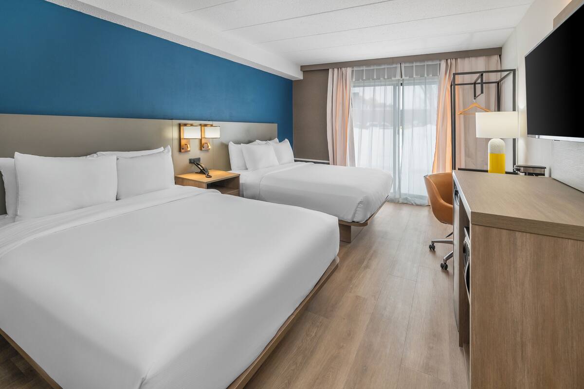 Une chambre d’hôtel avec deux lits, un bureau, une chaise, une lampe et une grande fenêtre avec des rideaux.