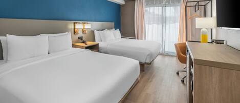 Habitación de hotel con dos camas, un escritorio, una silla, una lámpara y un amplio ventanal con cortinas.