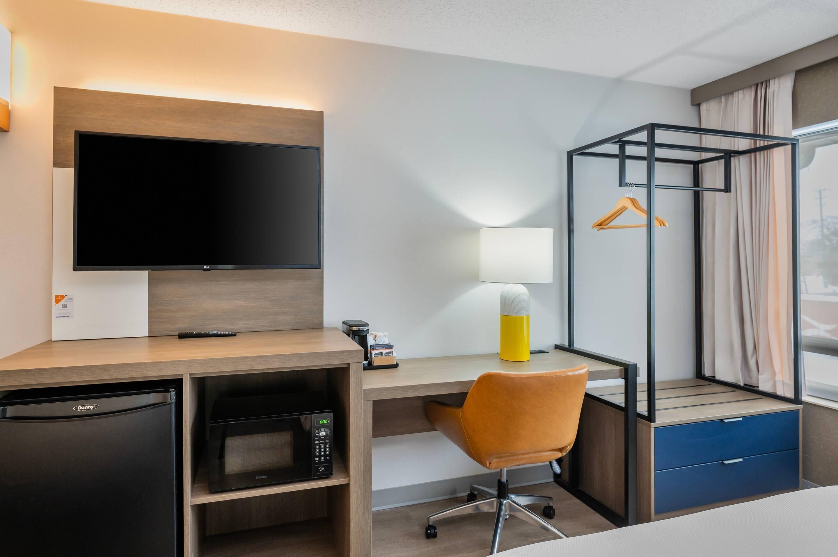 A hotel room with a flat-screen TV, a desk with a chair, a microwave, a mini-fridge, a hanging clothes rack, and a window with curtains.