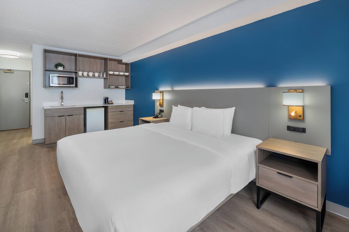 Une chambre d’hôtel moderne dotée d’un grand lit, d’une table de chevet, d’une petite commode, d’un four à micro-ondes encastré et d’une petite kitchenette.