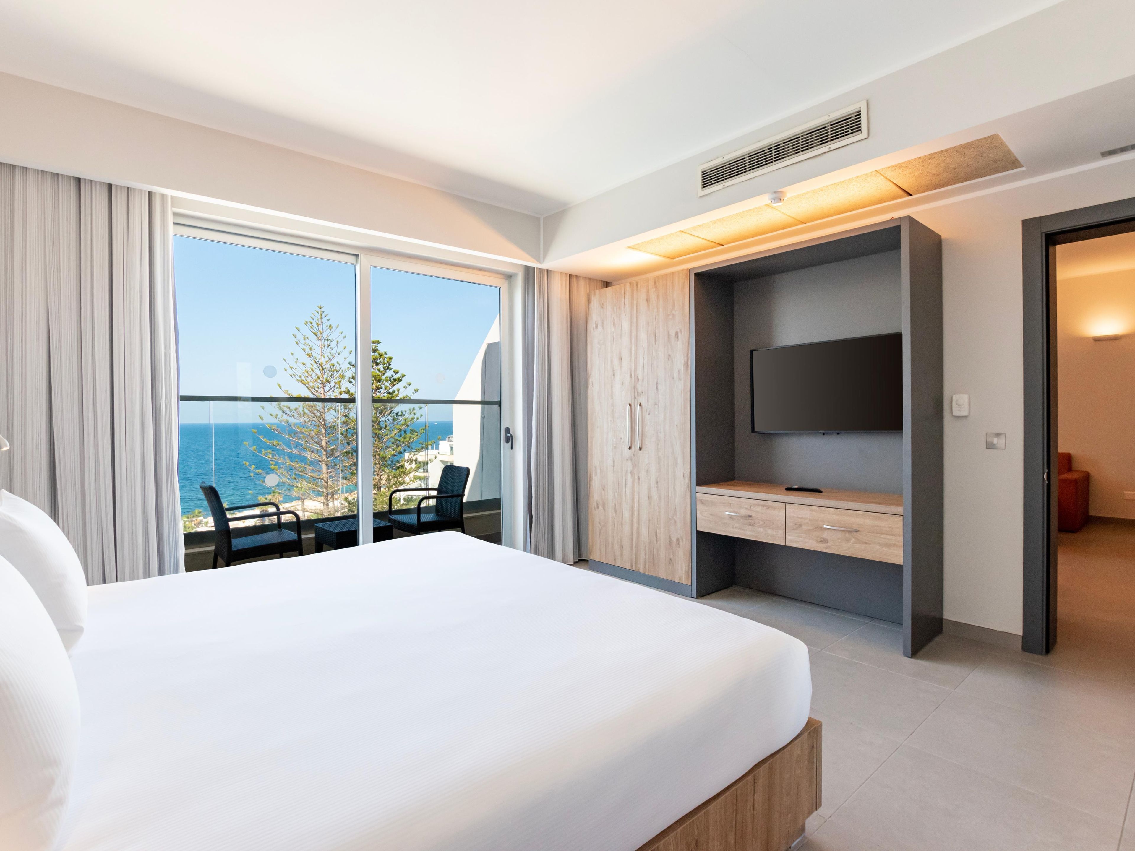 Ein Hotelzimmer mit einem großen Bett, einem Fernseher, einem Schreibtisch und einem Balkon mit Meerblick.
