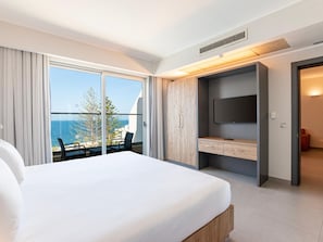 A hotel room with a large bed, a TV, a desk, and a balcony with a view of the sea.