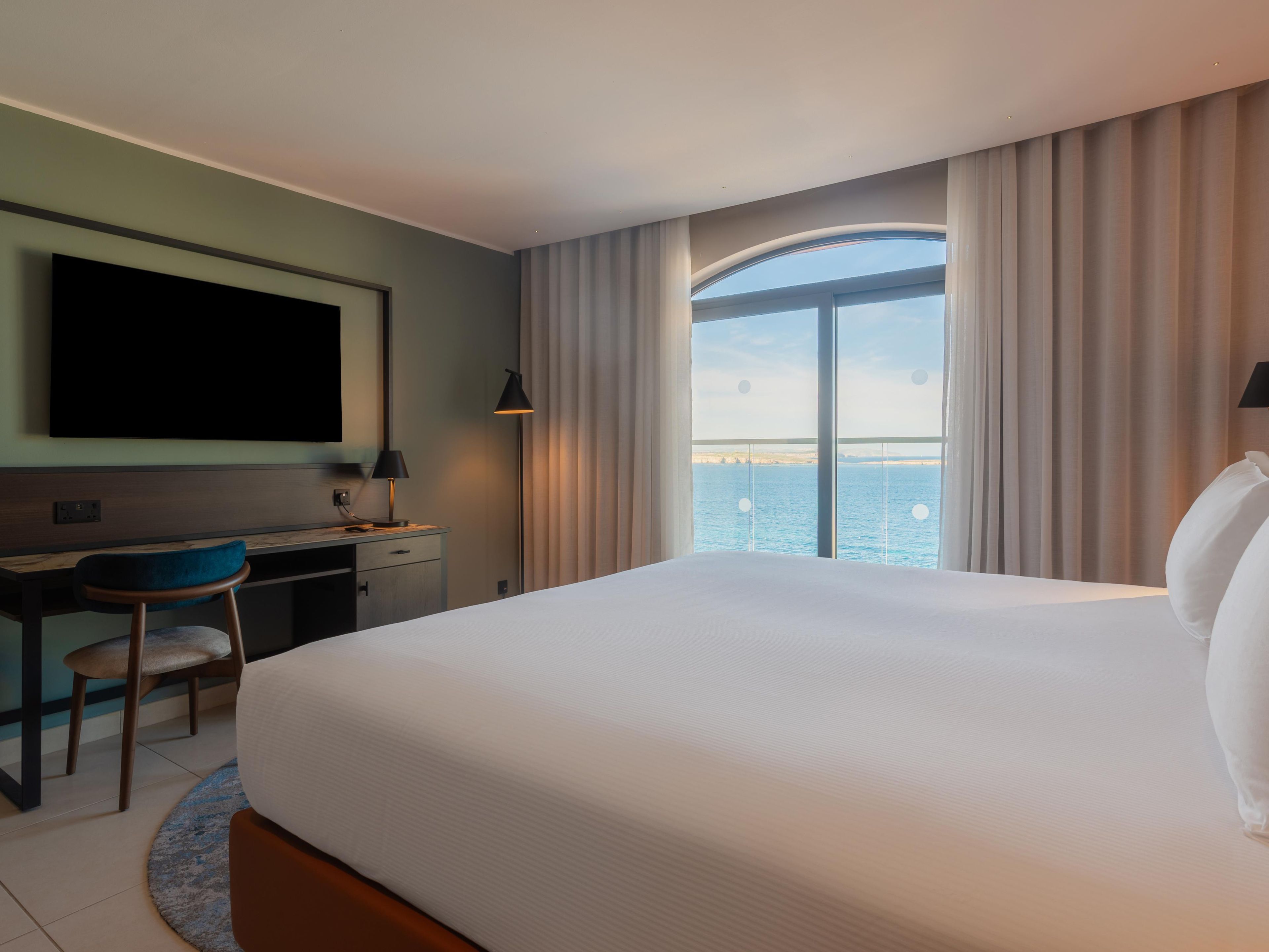 A hotel room with a bed, a desk with a chair, a TV, and a view of the sea.
