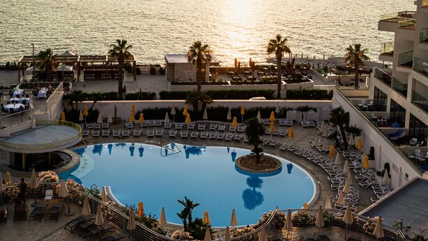 Piscina de un complejo turístico con tumbonas y sombrillas, rodeada por un edificio con vistas al mar.