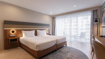 A hotel room with a double bed, bedside tables, a desk, and a large window with sheer curtains.